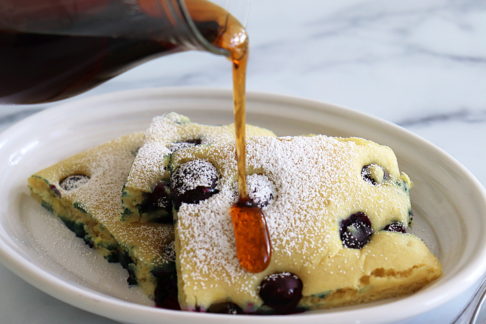 Pouring syrup on Powdered Sugar Vegan Lemon Blueberry Sheet Pan Pancakes